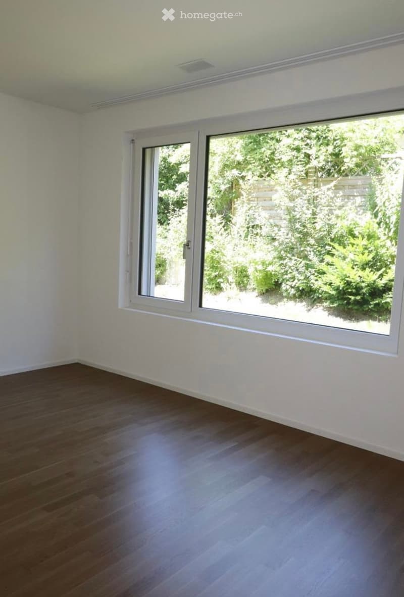 Empty room with wooden flooring and white walls, featuring large windows with white frames that open to a garden view.