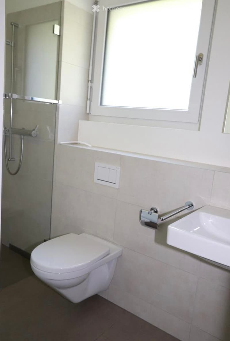 Modern bathroom with a toilet, sink, and glass-enclosed shower. Light-colored tiles and a window provide natural light.