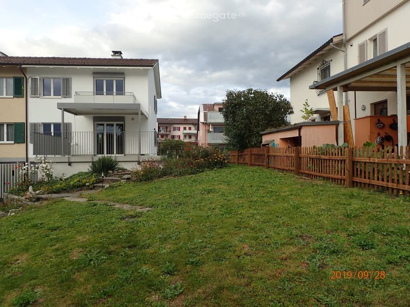Lush green lawn with a wooden fence, small garden beds, and a modern house with a balcony and large windows in the background.