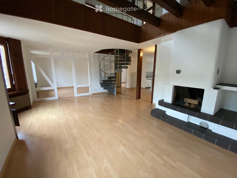 Empty room with light wooden flooring, white walls, and a fireplace. A spiral staircase leads to an upper level, and there are large windows and doors providing natural light.