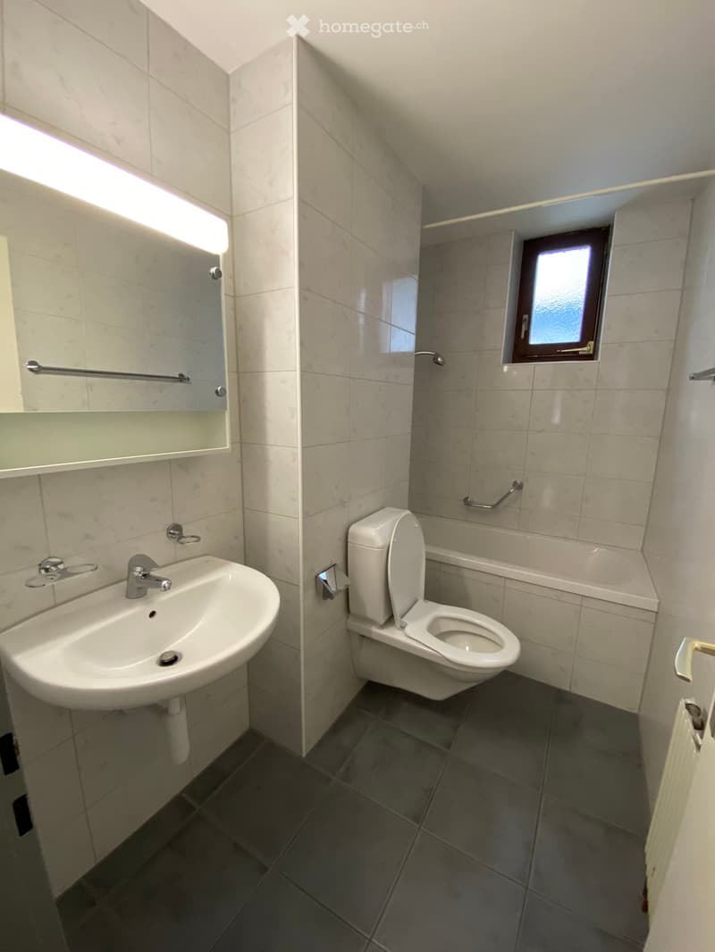 Modern bathroom with a white sink, toilet, and mirror. The walls are tiled in light beige, and the floor is dark grey tiles. A small window is visible in the shower area.