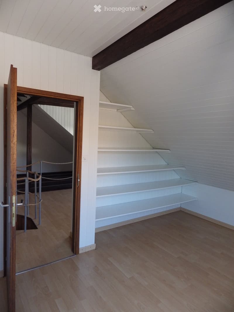 Empty room with wooden flooring, white paneled walls, built-in shelves, and a wooden door leading to a staircase.