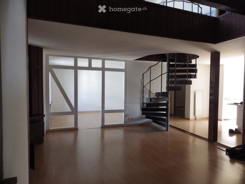 Open-plan unfurnished room with wooden flooring, large windows, and a modern spiral staircase leading to an upper level.