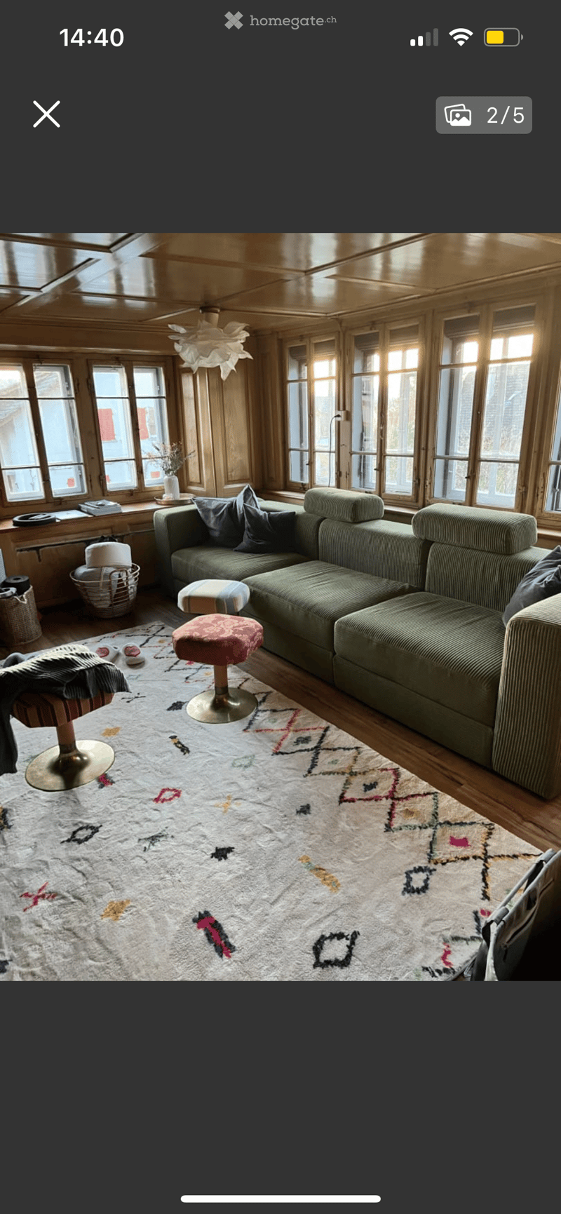 A cozy living room with wooden walls and large windows, featuring a green sectional sofa, colorful patterned rug, and various seating options including a stool and chair.
