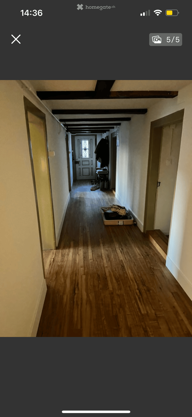 A long, narrow hallway with wooden flooring and white walls, featuring several doors and a staircase leading upwards.