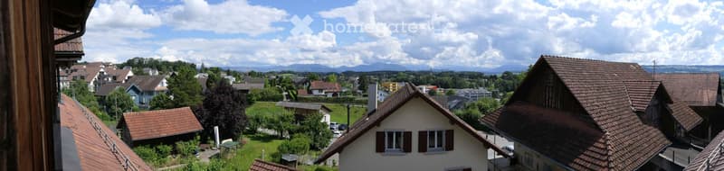 Balcony view overlooking a residential area with houses, trees, and distant mountains under a partly cloudy sky.