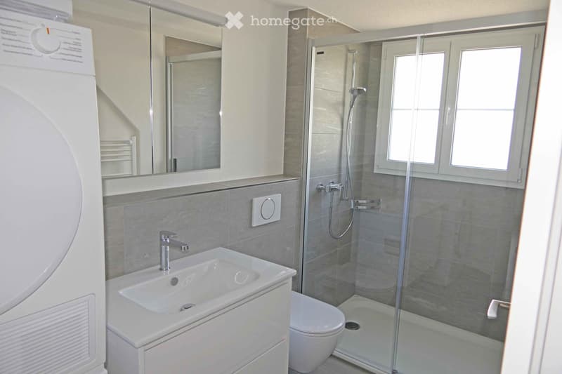 Modern bathroom with a white sink, chrome faucet, large mirror, toilet, and a glass-enclosed shower with a modern showerhead. The walls are tiled in a light grey color, and there is a window providing natural light.