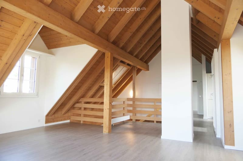 An empty attic space with exposed wooden beams, a wooden staircase leading up, and a large window letting in natural light. The floor is light-colored wood, and the walls are white.