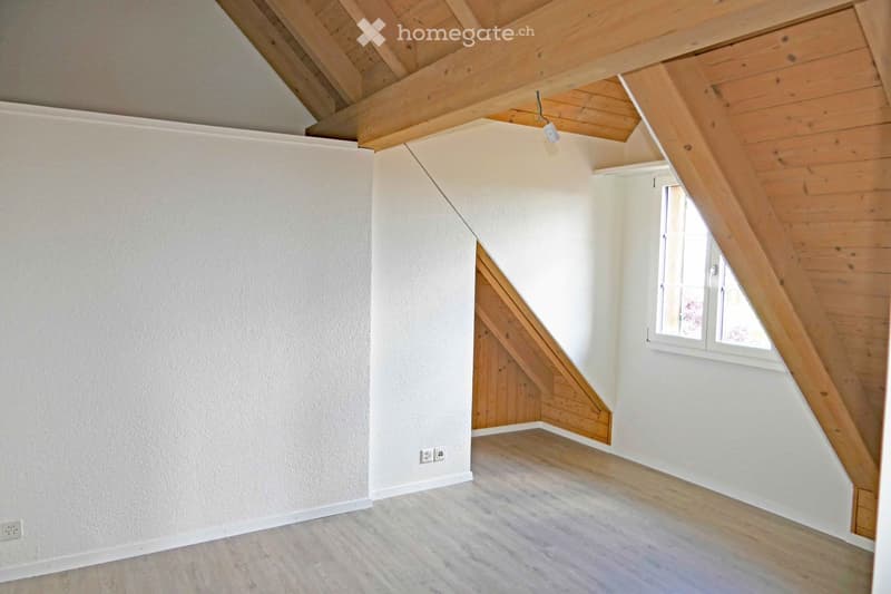 Empty attic room with wooden slanted ceilings, white walls, and a single window. The floor is light wood, and there are electrical outlets visible on the walls.