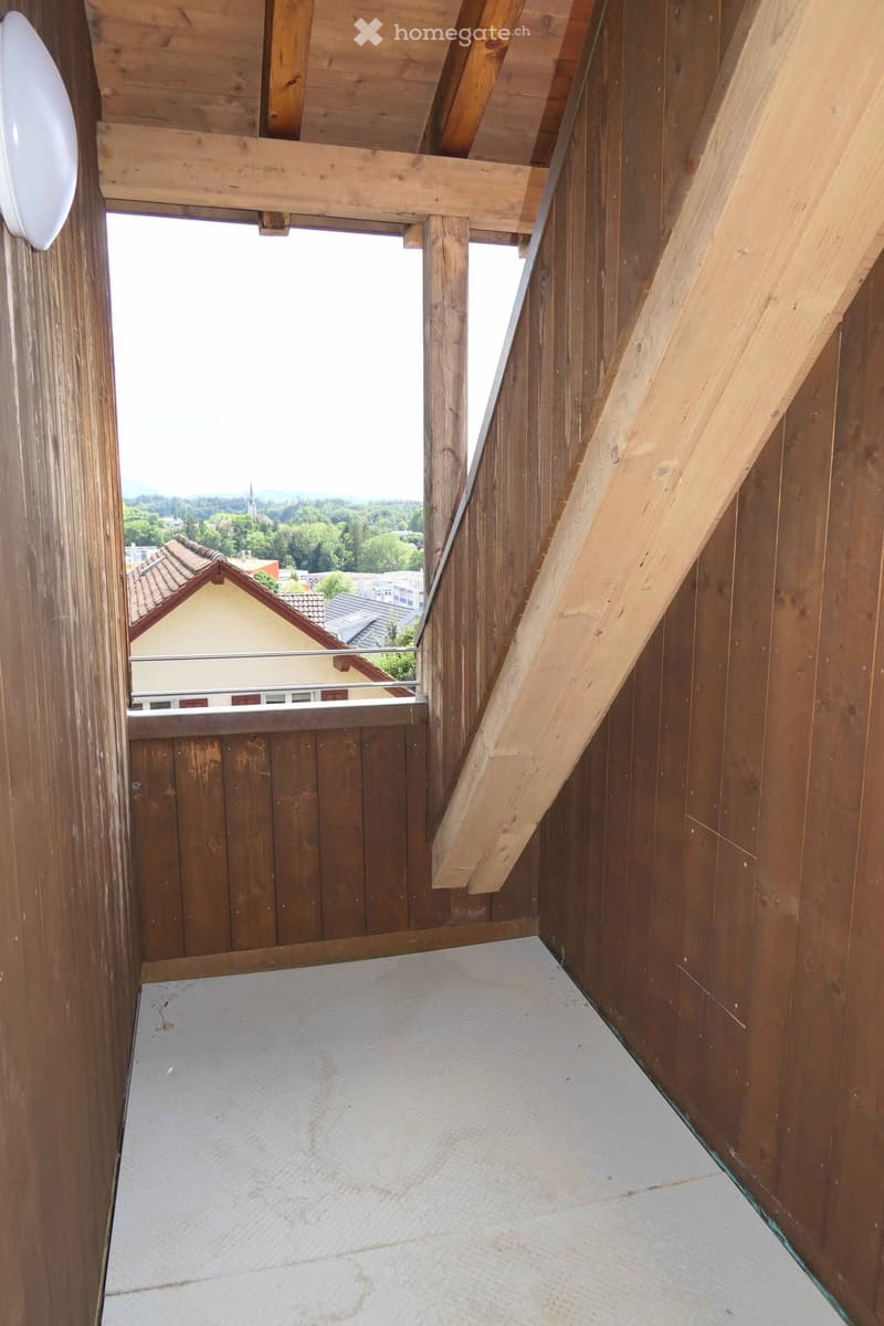 Wooden balcony with exposed beams, white tiled floor, and metal railing overlooking a town with houses and trees.