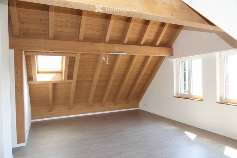 Empty attic room with wooden beams and exposed wooden ceiling, white walls, wooden floor, and multiple windows allowing natural light.