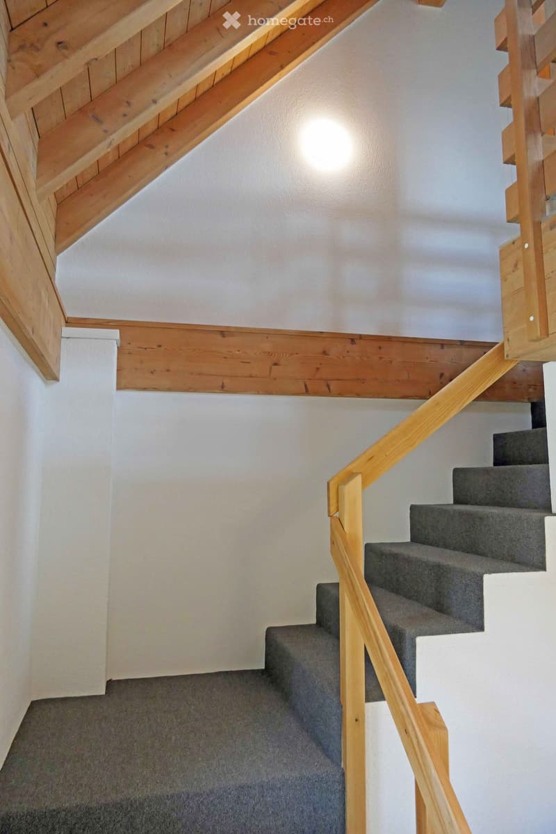 Wooden staircase with grey carpeted steps and a wooden handrail, set against white walls and a wooden ceiling with exposed beams.