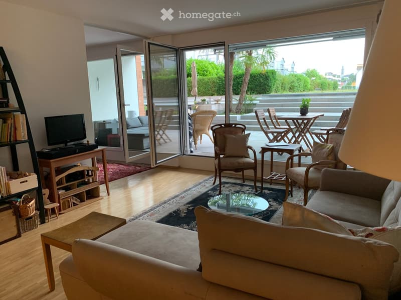 Bright living room with large sliding glass doors leading to a balcony, featuring a sofa, coffee table, chairs, and a television on a wooden stand.