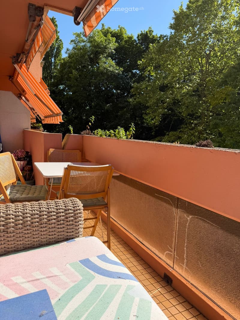 Balcony with a table, chairs, wicker basket, and ottoman, surrounded by greenery and trees.