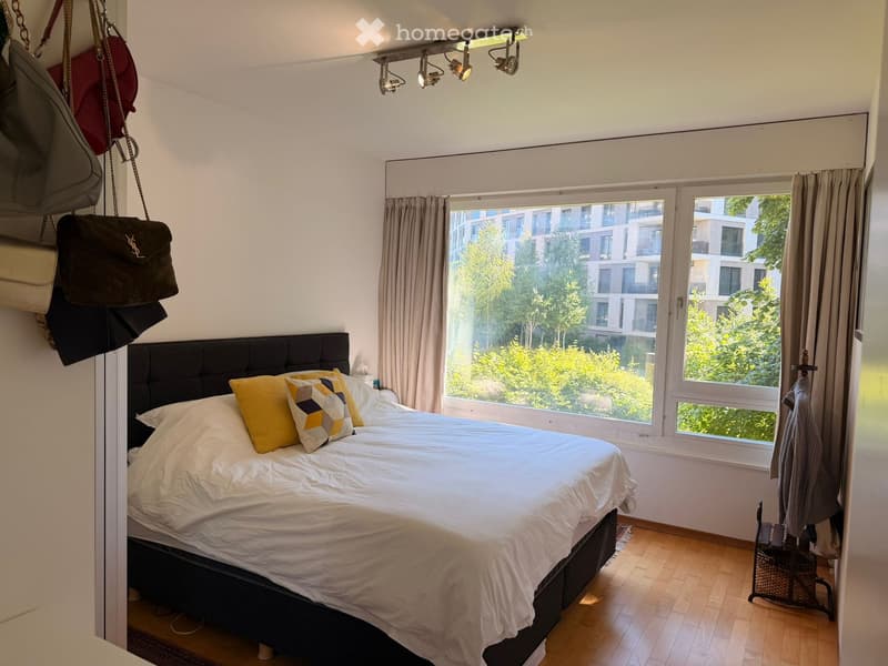 Modern bedroom with a large bed, yellow and grey patterned pillows, white bedding, wooden flooring, large windows with curtains, and several handbags hanging on the wall.