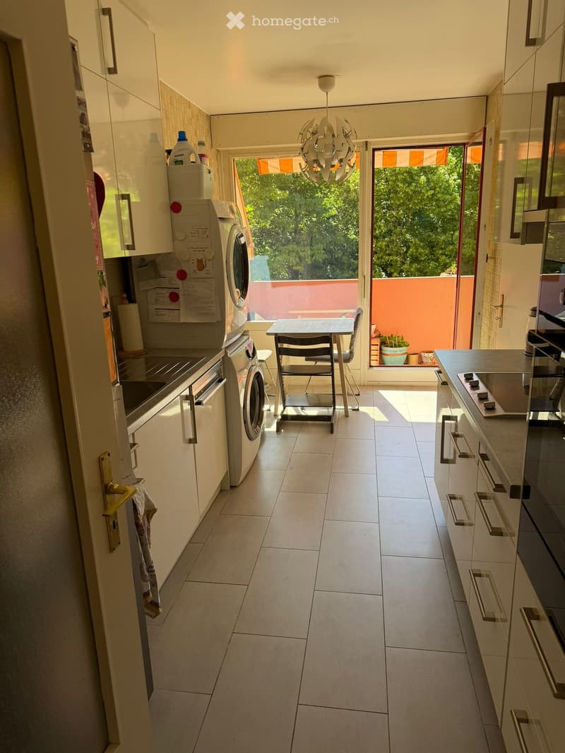 Modern kitchen with white cabinets, a washing machine, a small dining table with chairs, and a view to a balcony.