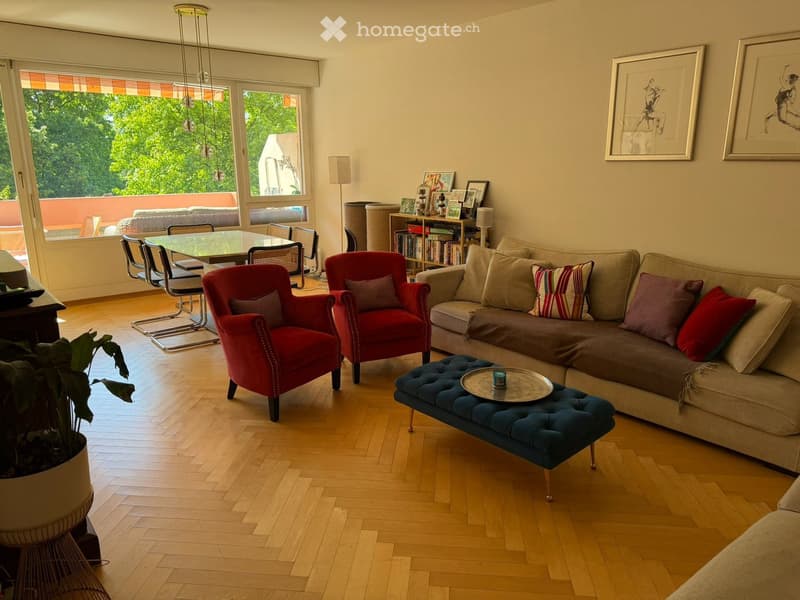 Bright living room with wooden flooring, large windows, a beige sofa with colorful cushions, a blue ottoman, red armchairs, a glass dining table with chairs, and a bookshelf with books and decorative items.