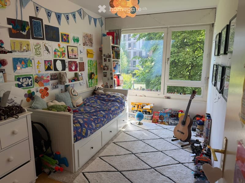 A cozy, personalized bedroom with a single bed, colorful bedding, a guitar, a bookshelf, a laptop, and various toys and drawings on the walls.