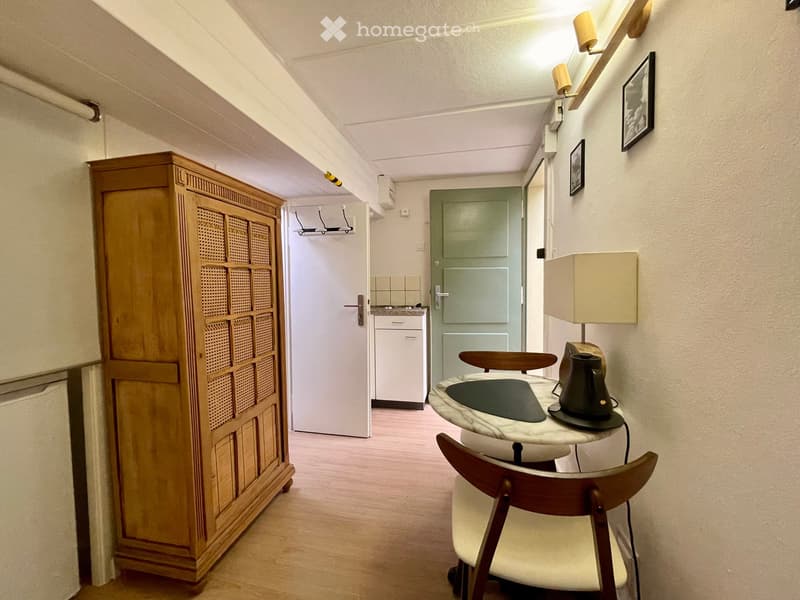 A small, furnished room with a wooden floor, a round table with a marble top, a chair, a lamp, a wooden cabinet, and a small kitchen area with a refrigerator and stove visible through an open door.