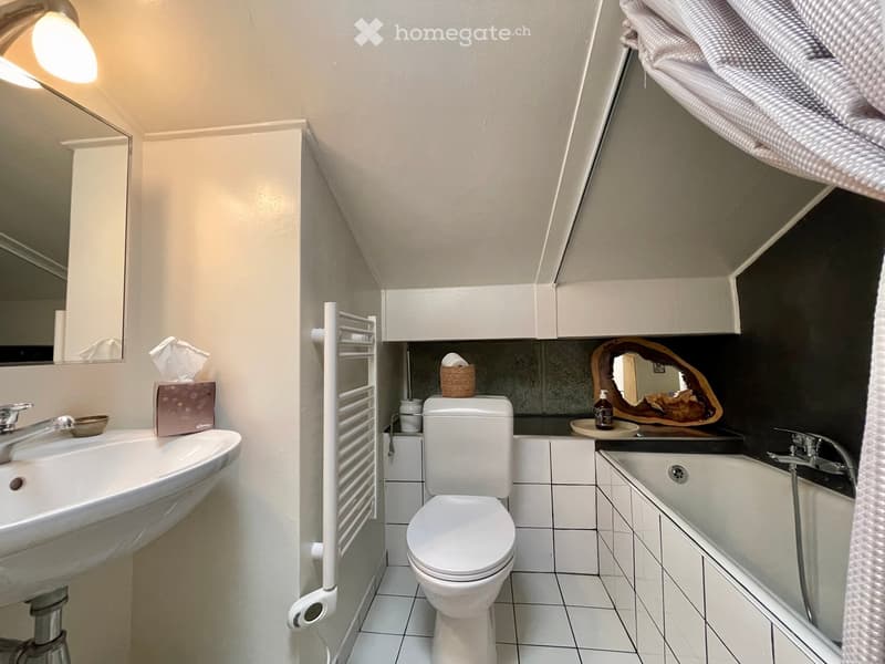Modern bathroom with white tiles, a sink, toilet, bathtub, and a towel rack. Features a decorative mirror and a small basket on the edge of the bathtub.