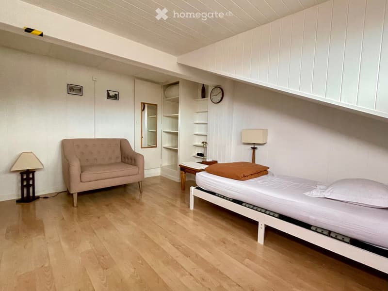 Cozy, well-lit room with a single bed, sofa, small table, and shelves. The room features wooden flooring and white walls.