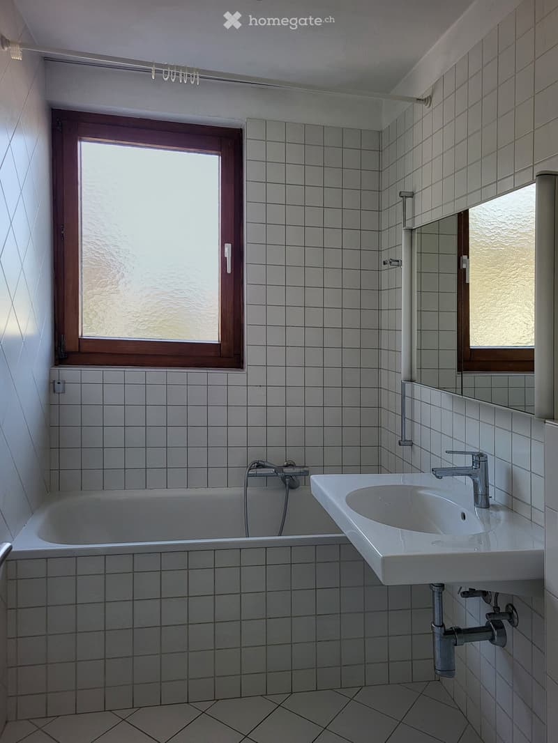 Bathroom with white tiled walls and floor, featuring a bathtub, a sink with a modern faucet, and a mirror above the sink. A window with wooden frame provides natural light.