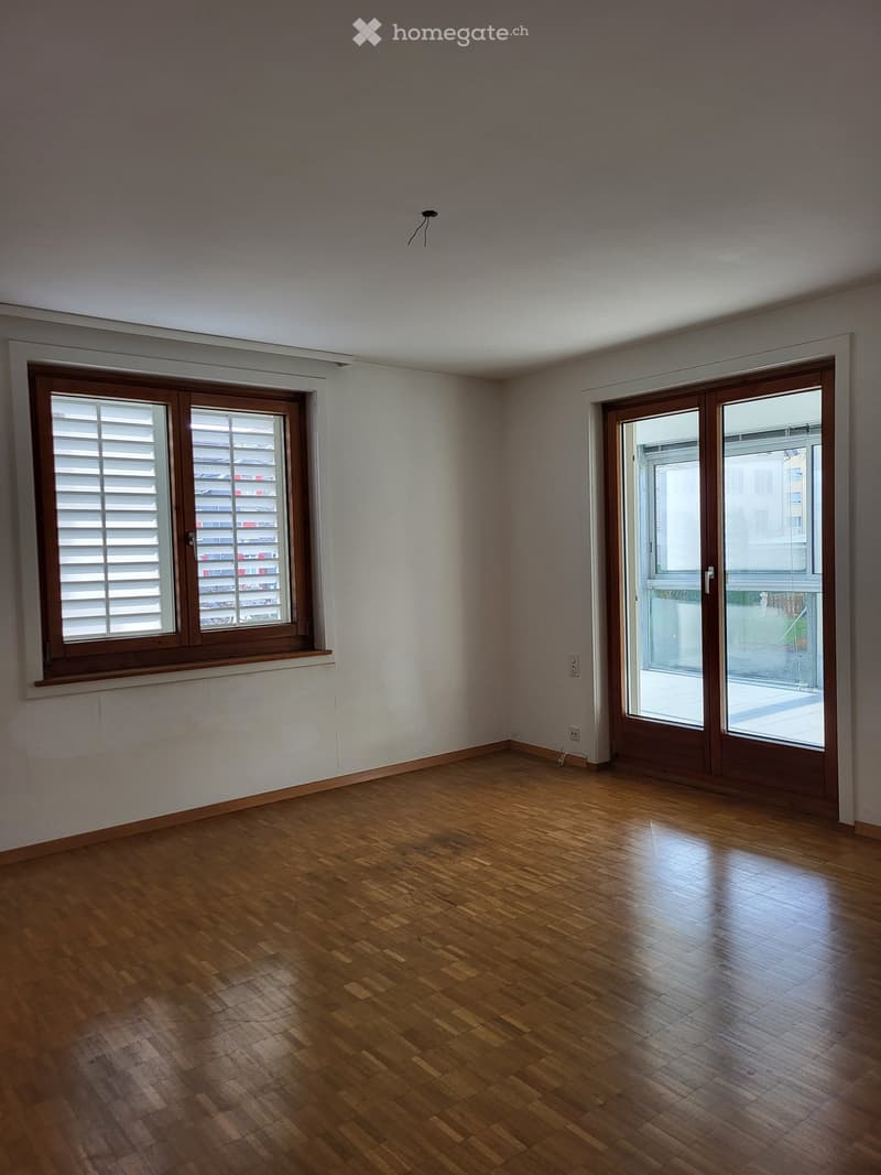 Empty room with wooden flooring, white walls, and large windows with wooden frames. A sliding glass door leads to a balcony area.