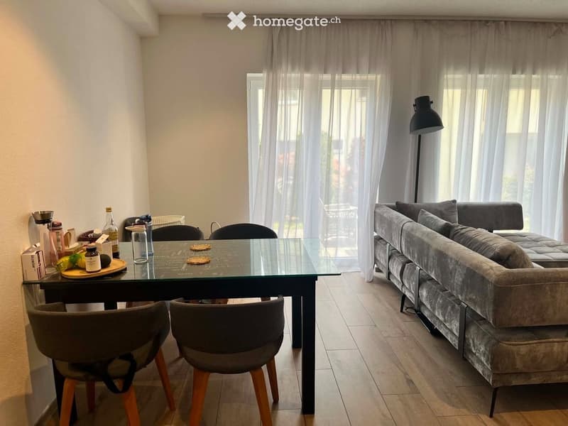 A modern living and dining area with a grey sectional sofa, a glass dining table with four chairs, and a floor lamp. The space features large windows with sheer curtains and a tiled floor.