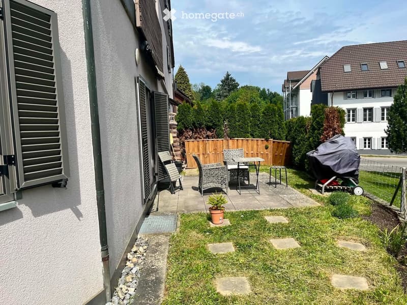 Small garden area with a table, chairs, a potted plant, and a covered lawn mower. The space is bordered by a wooden fence and a concrete pathway.