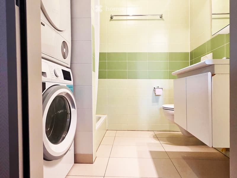 Modern bathroom with white tiles, a toilet, a sink with a mirror, a cabinet, and a towel rack. A washing machine is visible to the left.