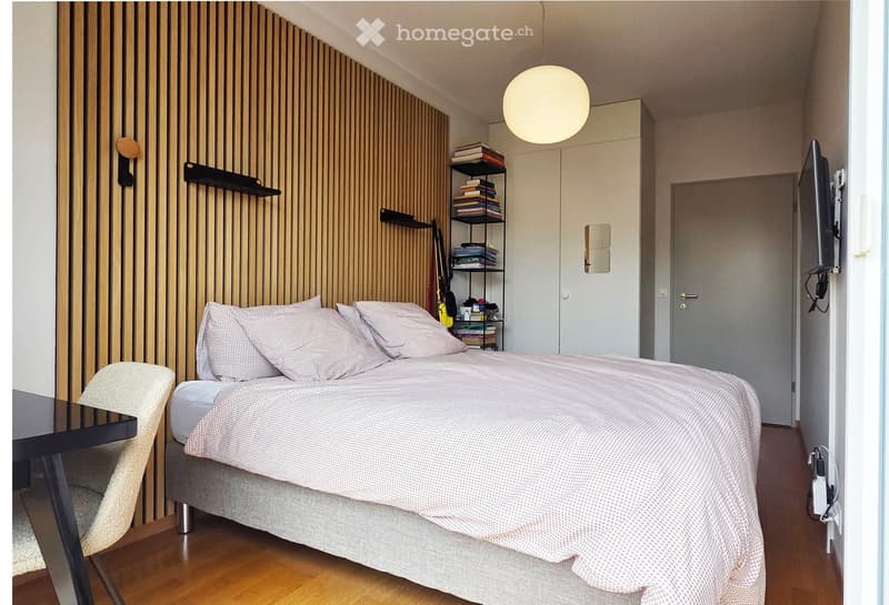 Modern bedroom with a wooden wall, a bed with a patterned duvet, a chair, a small table, and a shelf with books and decorative items.