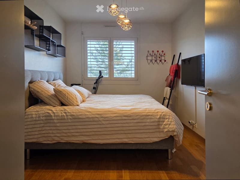 Modern bedroom with a large bed, striped bedding, wall-mounted shelves, a coat rack, and a television. The room features wooden flooring and large windows with blinds.