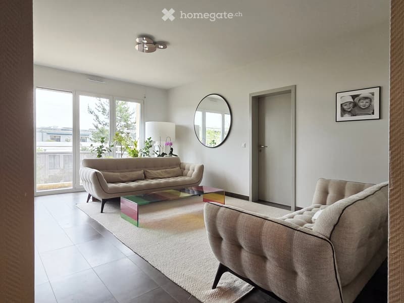 Modern living room with a beige sofa, coffee table, floor lamp, round mirror, and picture frame on the wall. Large windows and sliding doors lead to a balcony.