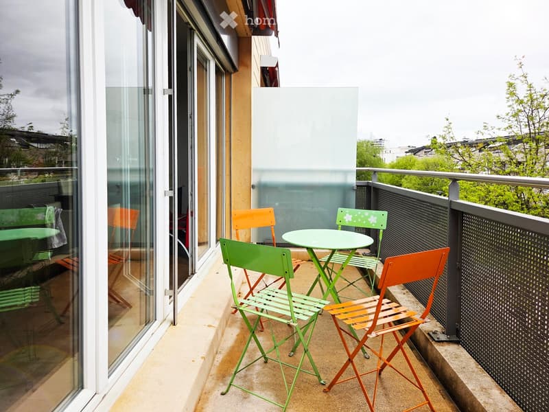 Balcony with a small round table and four colorful chairs, overlooking a cityscape with trees and buildings.