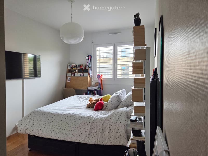 A well-lit bedroom with a large bed, pillows, and a patterned bedspread. A wooden desk with a chair is positioned near a window, and a bookshelf holds books and decorative items. A round pendant light hangs from the ceiling.