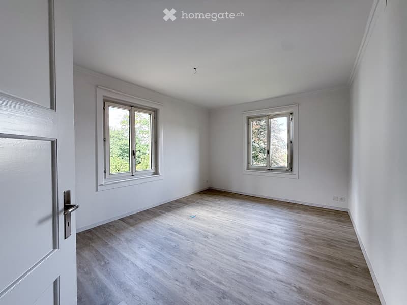 Empty room with white walls, light wooden flooring, and two windows with white frames. The room has a simple, clean design with no furniture or decorations.