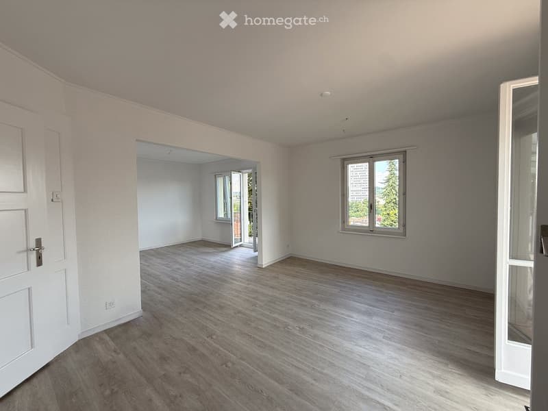 Empty room with light wooden flooring, white walls, and large windows offering a view of trees and buildings. The room features a door and an open doorway leading to another space.
