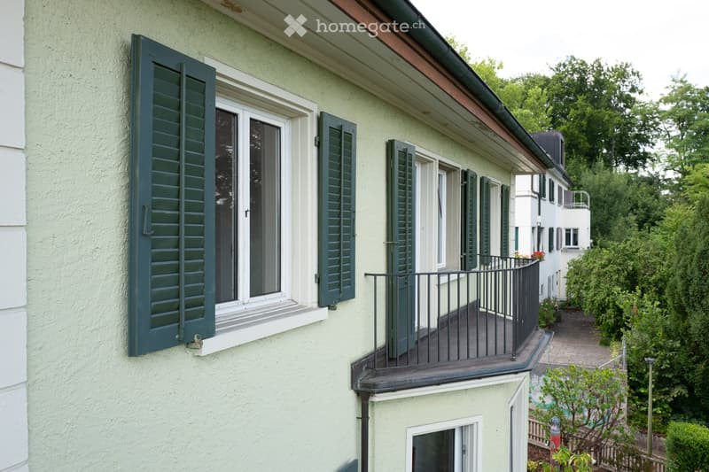 A two-story building with light green exterior walls, dark green shutters, and a small balcony with a black metal railing. The building features multiple windows and a sloped roof with a visible gutter.