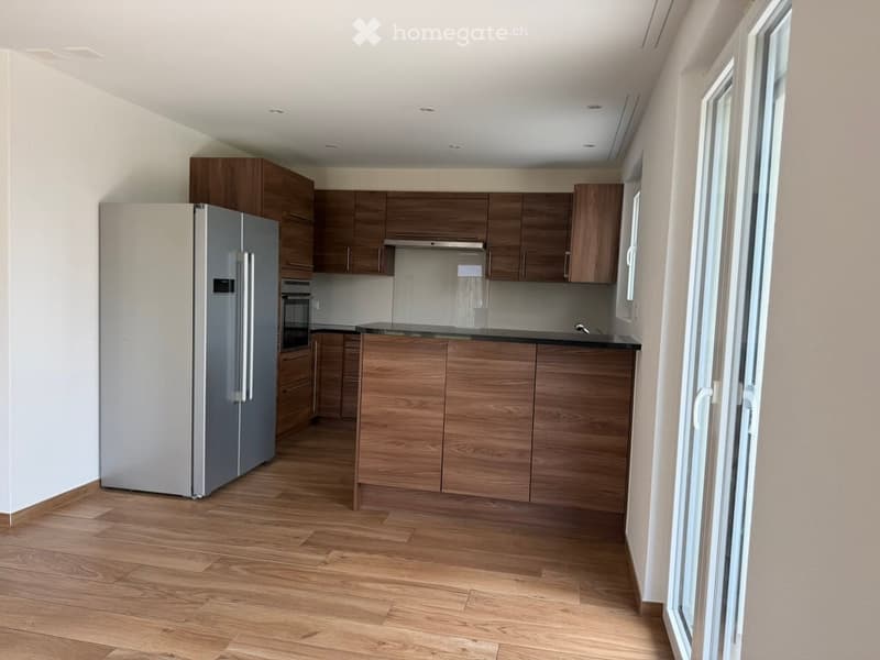 Modern kitchen with wooden cabinets, stainless steel refrigerator, and a central island. The space features light wooden flooring and large windows.