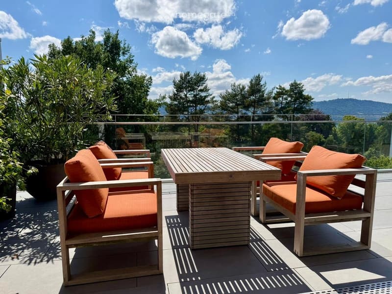 Balcony with wooden table and chairs, orange cushions, surrounded by greenery and a scenic view.