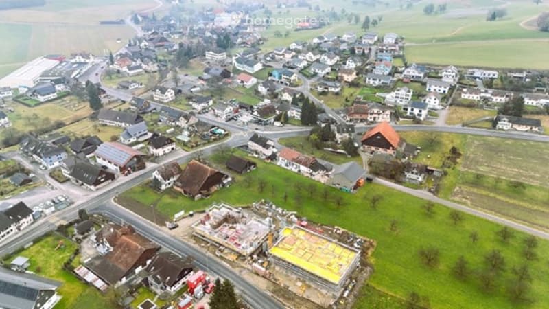 Aerial view of a rural village with scattered houses, roads, and green fields. Some buildings are under construction.