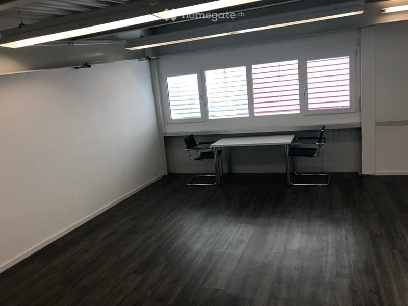 A small, unfurnished room with a dark wooden floor, white walls, and large windows with blinds. A small white table and two black chairs are placed near the windows.