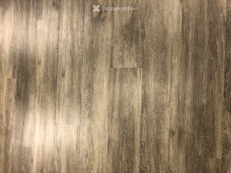 Close-up view of a wooden floor with a natural, textured finish. The floorboards are wide and have a warm, earthy tone with visible grain patterns.