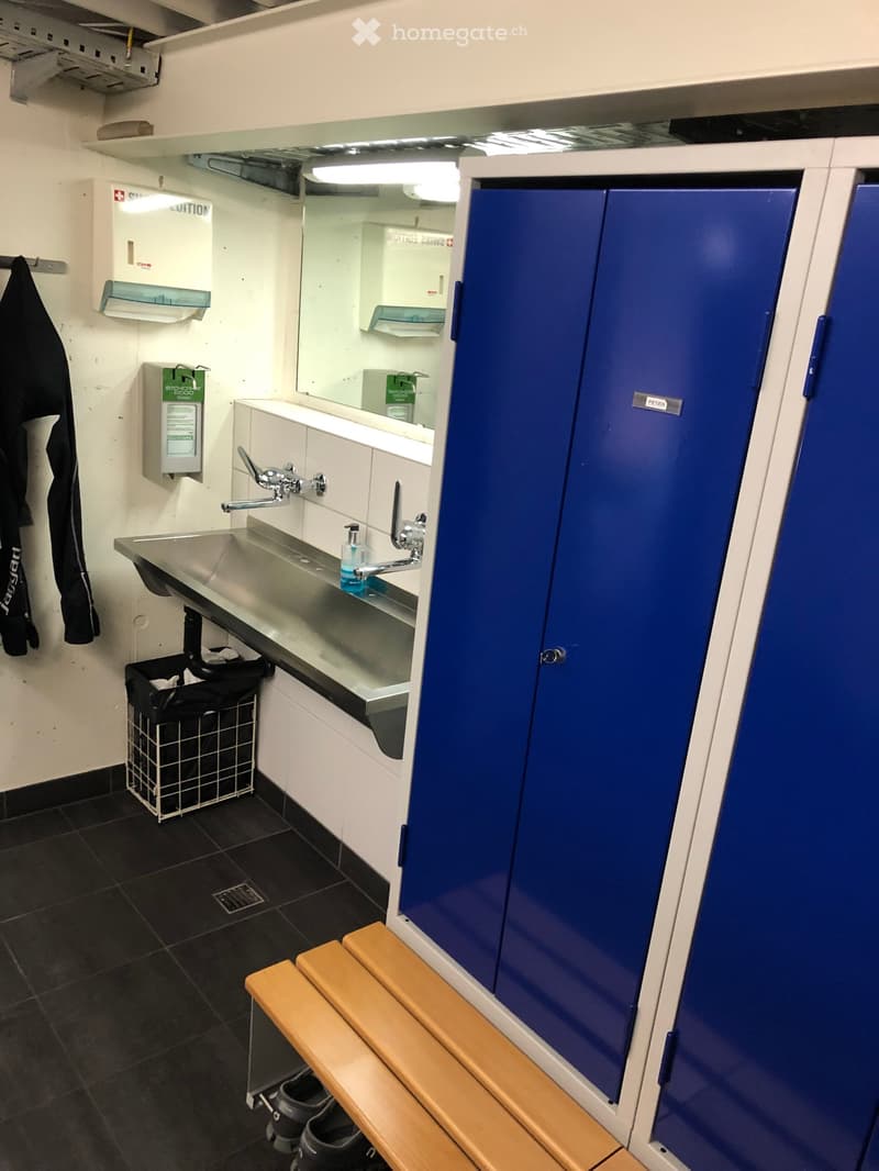 A small, clean bathroom with a stainless steel sink, mirror, wooden bench, blue lockers, and a coat rack with a jacket hanging on it.