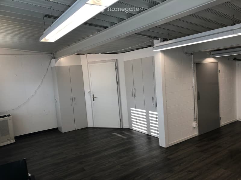 Empty room with wooden flooring, white walls, and several doors. Exposed ceiling beams and lighting fixtures are visible.