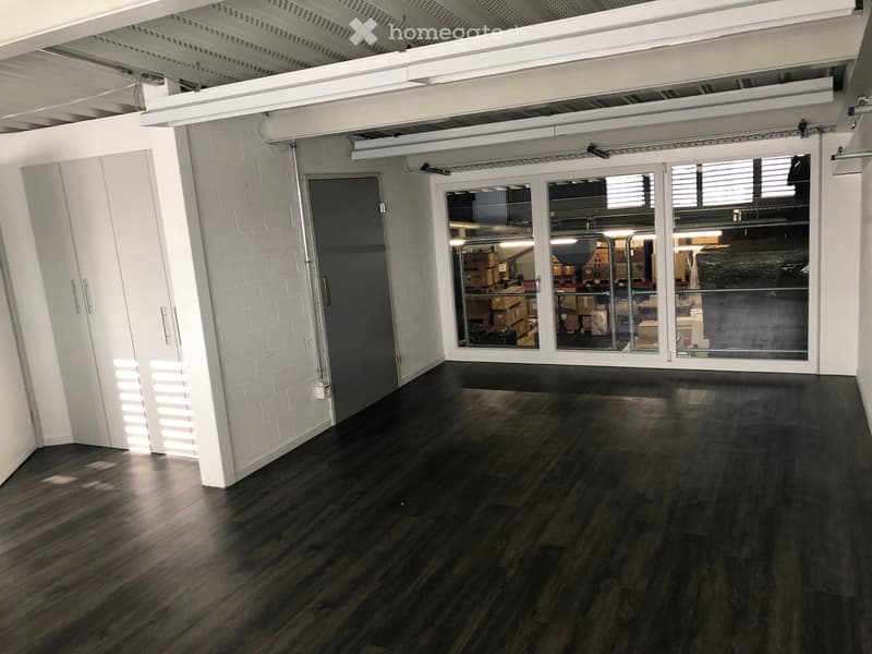 Empty room with dark wooden flooring, white brick walls, large glass doors, and exposed white ceiling beams.