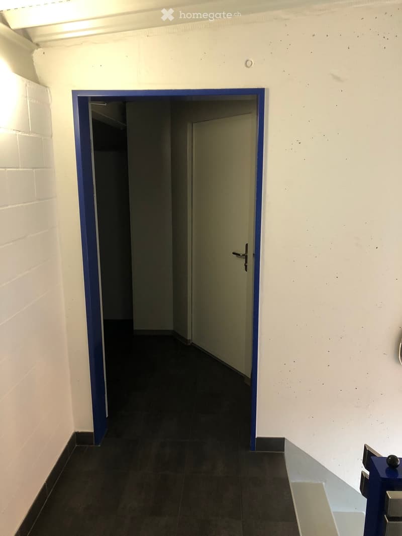 A narrow, well-lit corridor with white walls, dark tiled flooring, and a blue-framed doorway leading to another room.