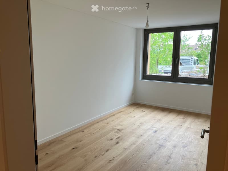 Empty room with light wooden flooring and white walls, featuring a large window with a view of greenery and a parked vehicle outside.