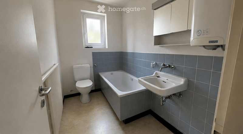Bathroom with a toilet, bathtub, sink, mirror, and cabinet. Blue tiles on lower walls, white upper walls, and a window providing natural light.
