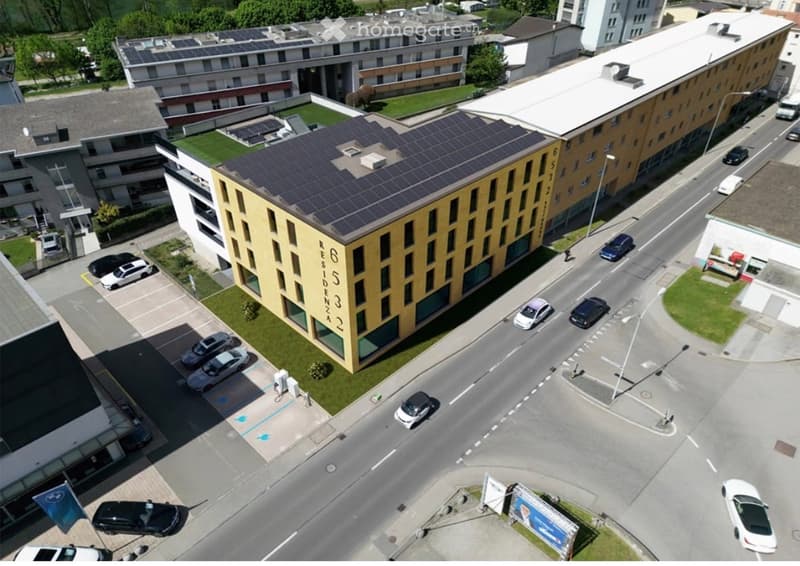 Aerial view of a modern multi-story residential building with solar panels on the roof, surrounded by parked cars and greenery.